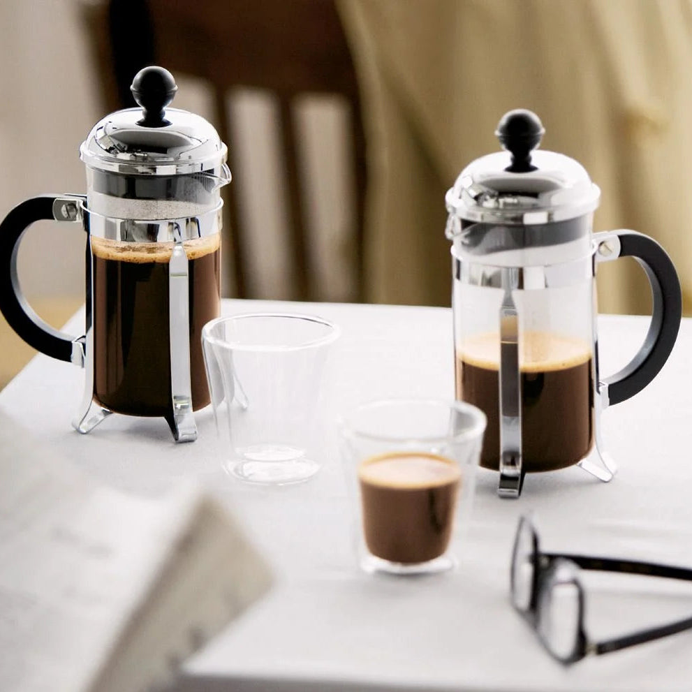 Two Bodum Chambord French Press coffee makers with coffee in them, one with a lid on and one with a lid off showing the plunger. They are placed on a table with a cup of coffee and a newspaper in the background.